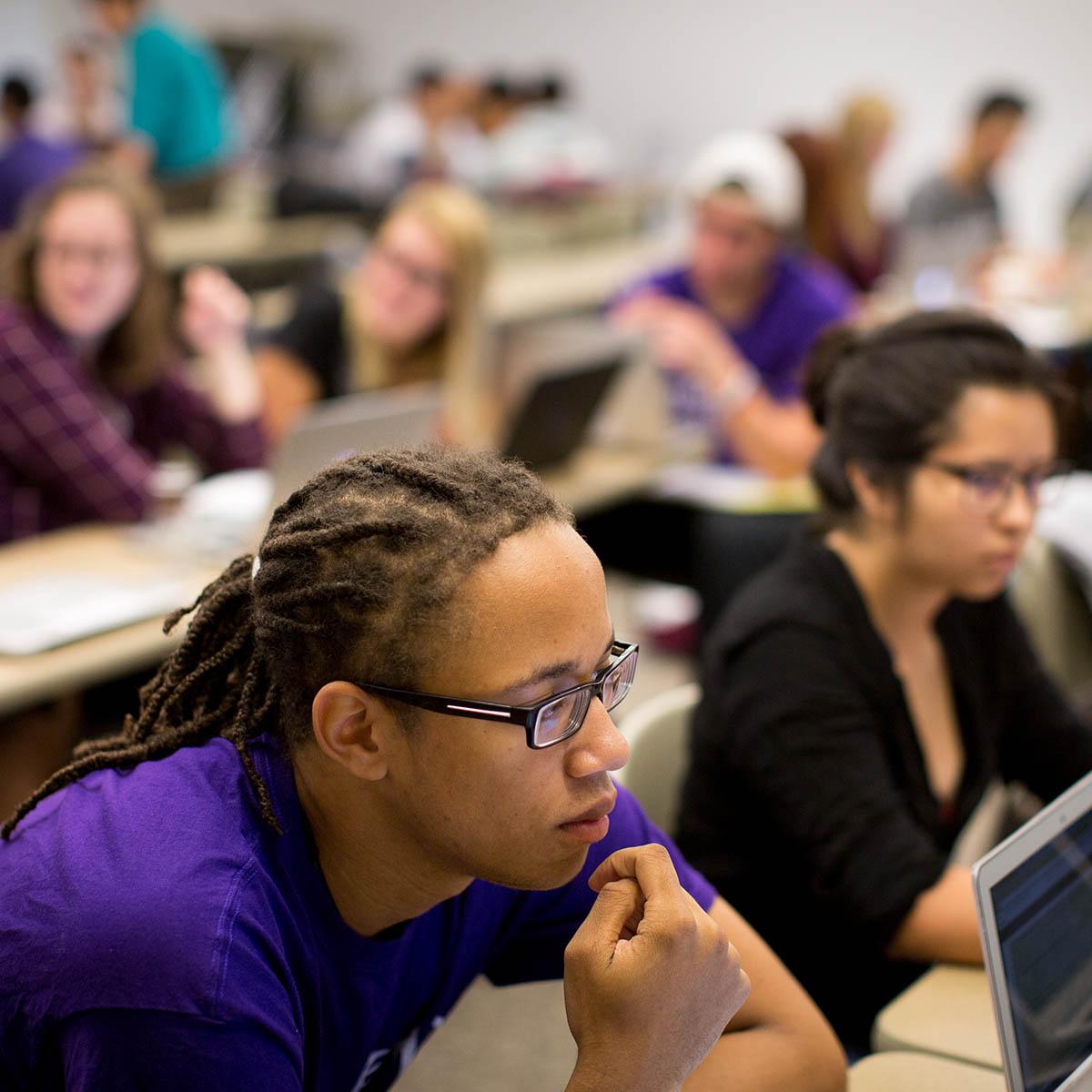 Photo of students listening in a classroom, with a young Black man with dreads, glasses, and a purple Chatham shirt in the foreground