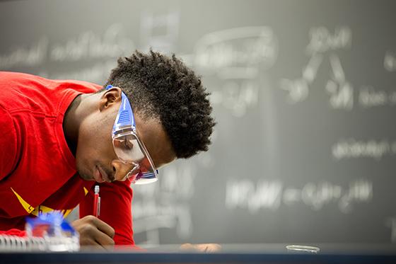 Photo of a Chatham University student wearing a red shirt and protective goggles in a lab, writing with a pencil. 